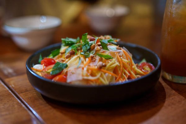 istockphoto-2182655497-612×612 The vibrant salad, which features shredded green papaya, mango, carrots, tomatoes, peanuts, and fresh herbs, is served in a dark bowl on a wooden table.